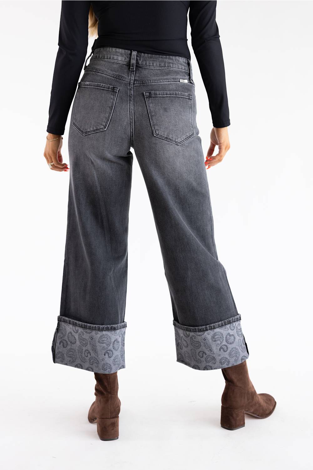 A person shown from the back wearing dark gray wide-leg cropped jeans with a paisley-patterned cuff, a black long-sleeve top, and brown ankle boots against a plain white studio background.