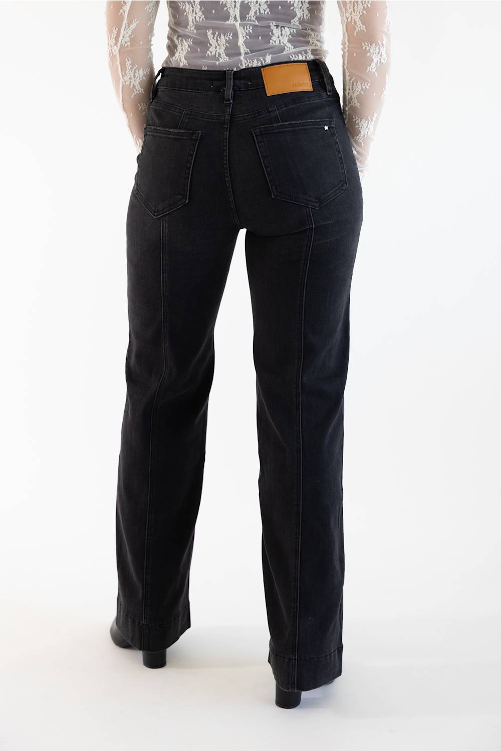 Rear view of a person wearing black wide-leg jeans with a tan leather patch at the back waistband, paired with a sheer patterned top and black heels, against a plain white studio background.