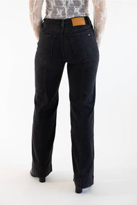 Rear view of a person wearing black wide-leg jeans with a tan leather patch at the back waistband, paired with a sheer patterned top and black heels, against a plain white studio background.