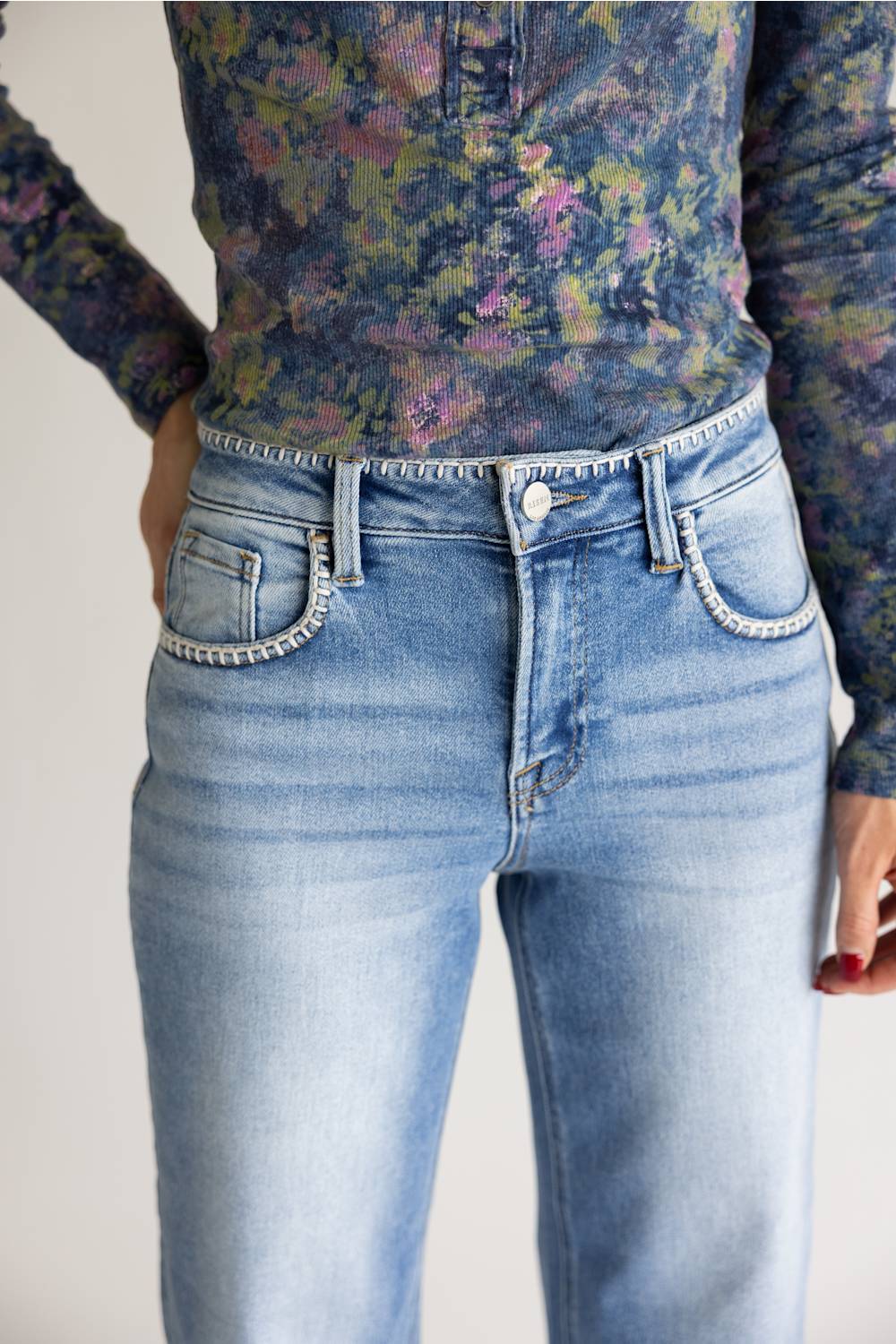 A person wearing light-wash blue jeans with decorative whipstitch trim along the pocket edges, paired with a floral long-sleeve top. The photo is cropped from the chest down against a plain light background.