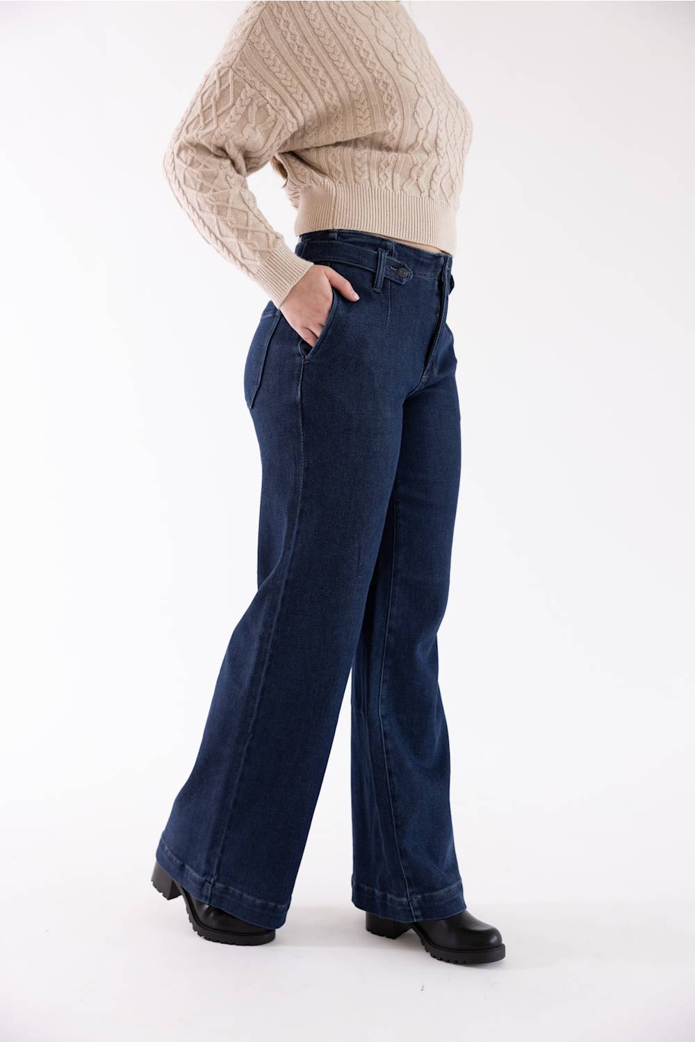A person wearing dark blue wide-leg jeans, a beige cable-knit sweater, and black shoes, shown from the chest down in a studio setting.