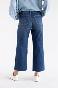 Rear view of a person wearing blue wide-leg cropped jeans with a light blue sweater and white sneakers against a plain white studio background.