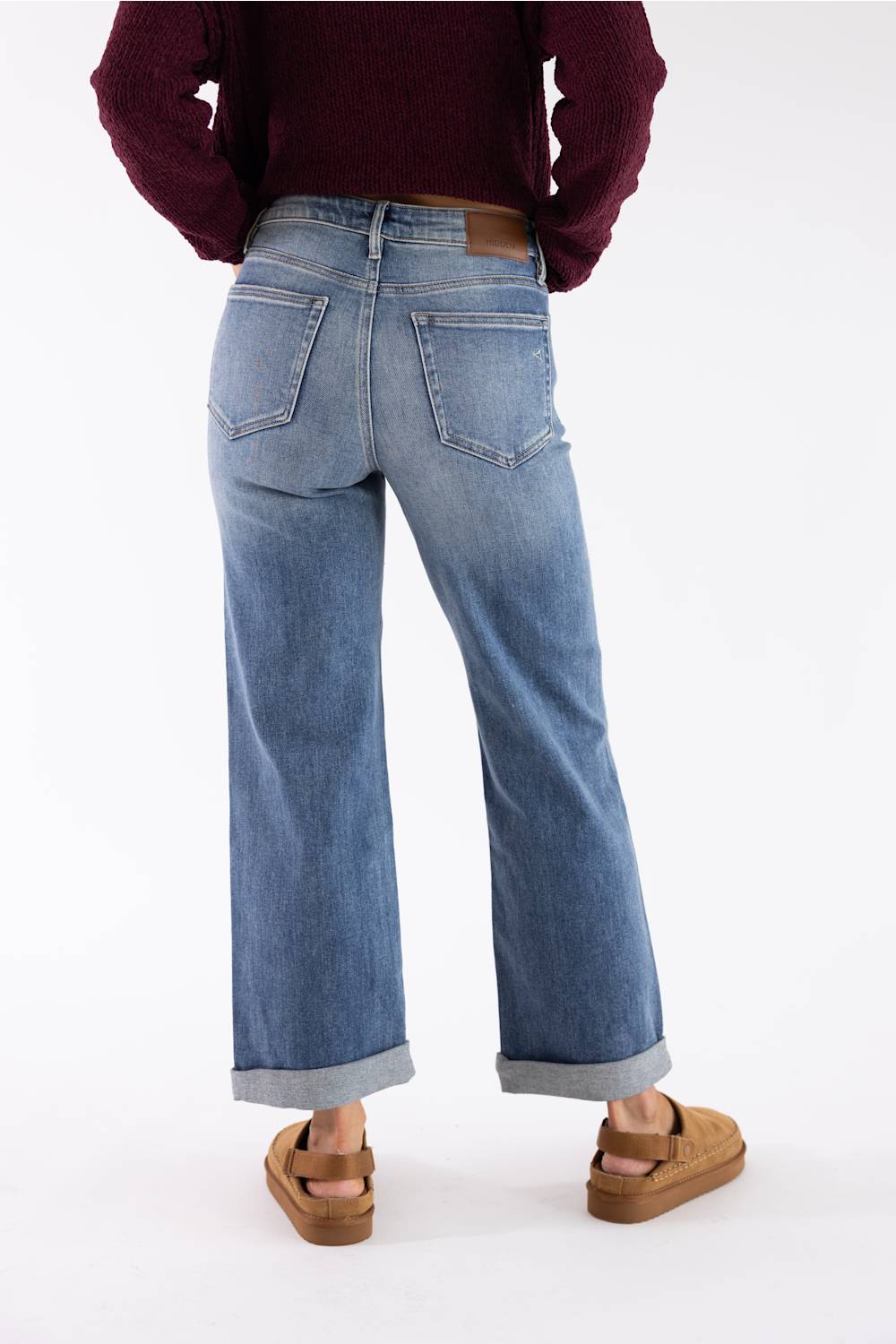A person shown from the back wearing light-wash blue wide-leg jeans with rolled cuffs, a burgundy sweater, and tan sandals against a plain white studio background.