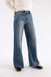 A person wearing blue wide-leg denim jeans with a cream ribbed sweater and black shoes, shown from the chest down against a plain light gray studio background.