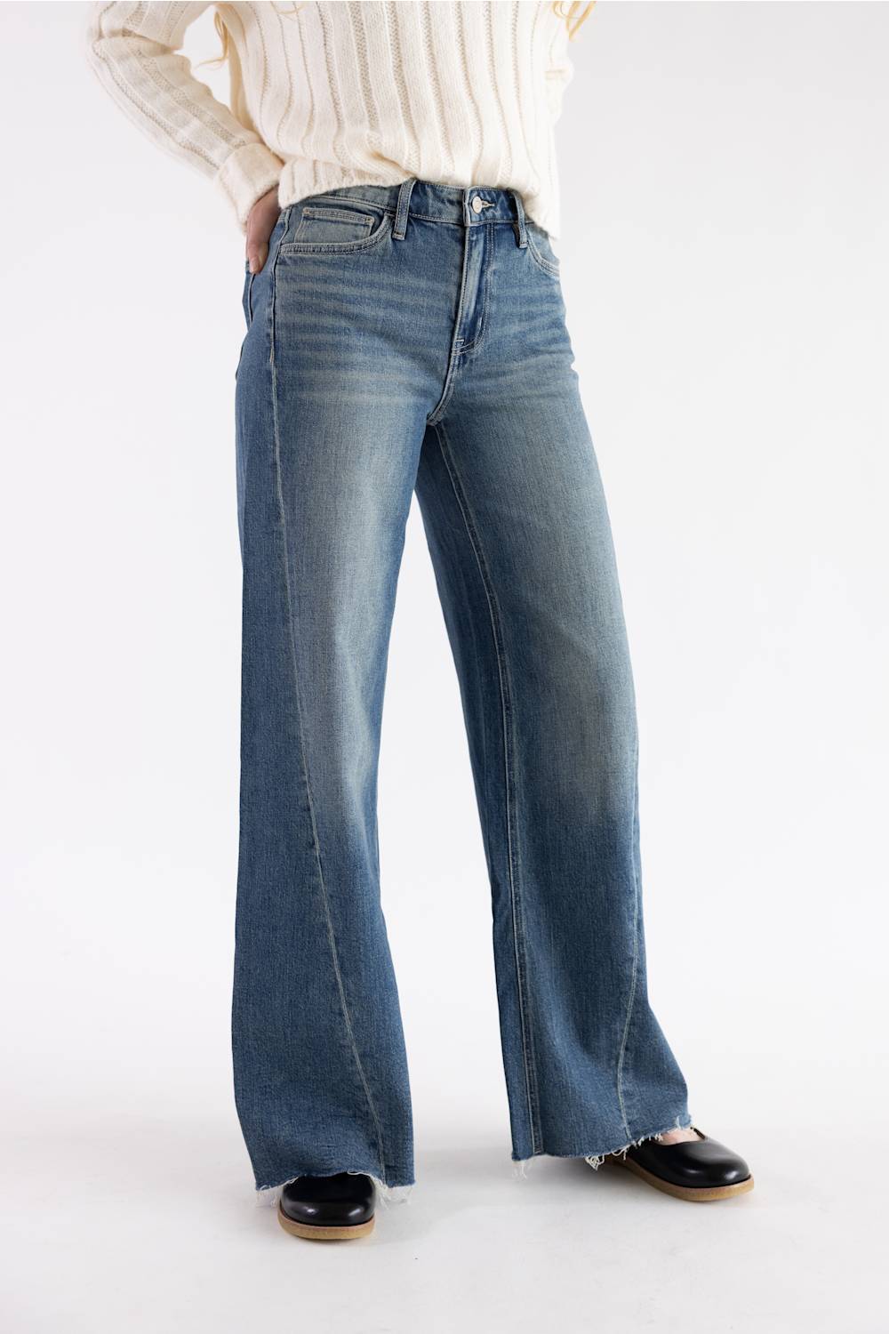 A person wearing blue wide-leg denim jeans with a cream ribbed sweater and black shoes, shown from the chest down against a plain light gray studio background.