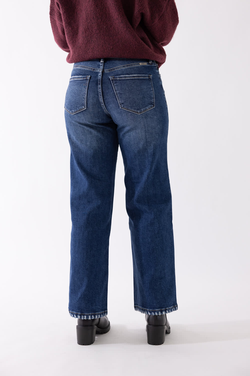 Back view of a model wearing KanCan dark wash, high-rise wide-leg jeans, styled with black boots and a burgundy sweater against a plain backdrop.