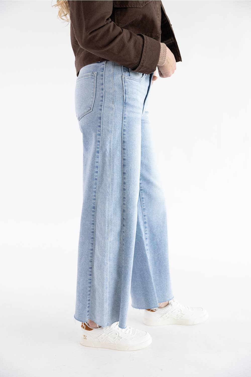 A person wearing light-wash wide-leg denim jeans, a dark brown jacket, and white sneakers, shown from the waist down against a plain white studio background.