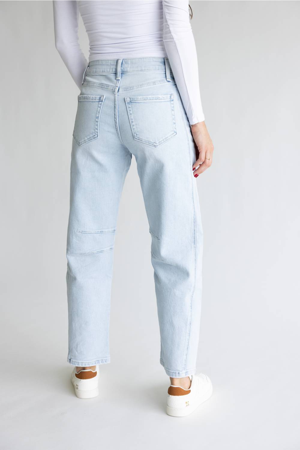 A person shown from the back wearing light-wash light blue jeans, a white long-sleeve top, and white sneakers against a plain light gray studio background.