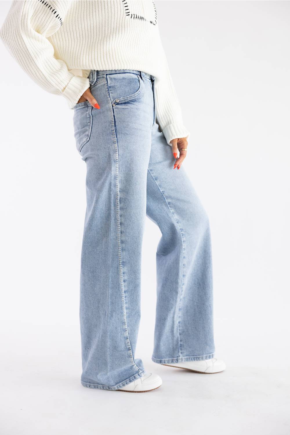 A person wearing light-wash wide-leg denim jeans with a cream ribbed sweater and white shoes, standing against a plain white studio background.