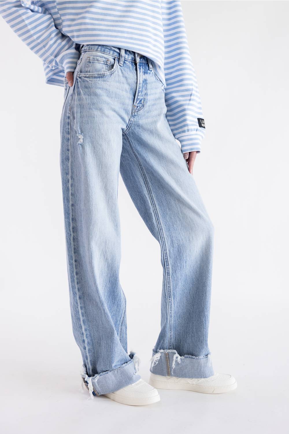 A person wearing light-wash wide-leg denim jeans with rolled cuffs, paired with a blue-and-white striped long-sleeve top and white sneakers, photographed against a clean white studio background.