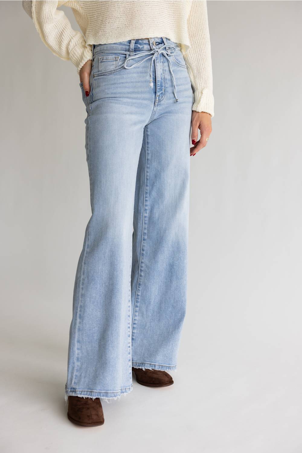 A person wearing light-wash blue wide-leg jeans with a drawstring waist, paired with a cream ribbed sweater and brown shoes, photographed against a plain light gray studio background.