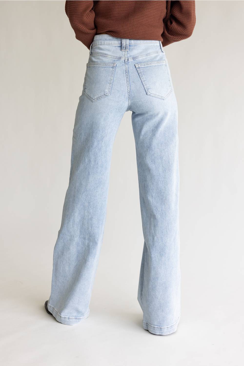 A person shown from the back wearing very light blue wide-leg jeans and a brown ribbed sweater, standing against a plain light beige studio background.