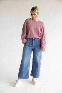 A woman modeling a dusty rose long-sleeve cropped sweatshirt and wide-leg blue jeans with white slip-on shoes, standing against a plain white studio background.