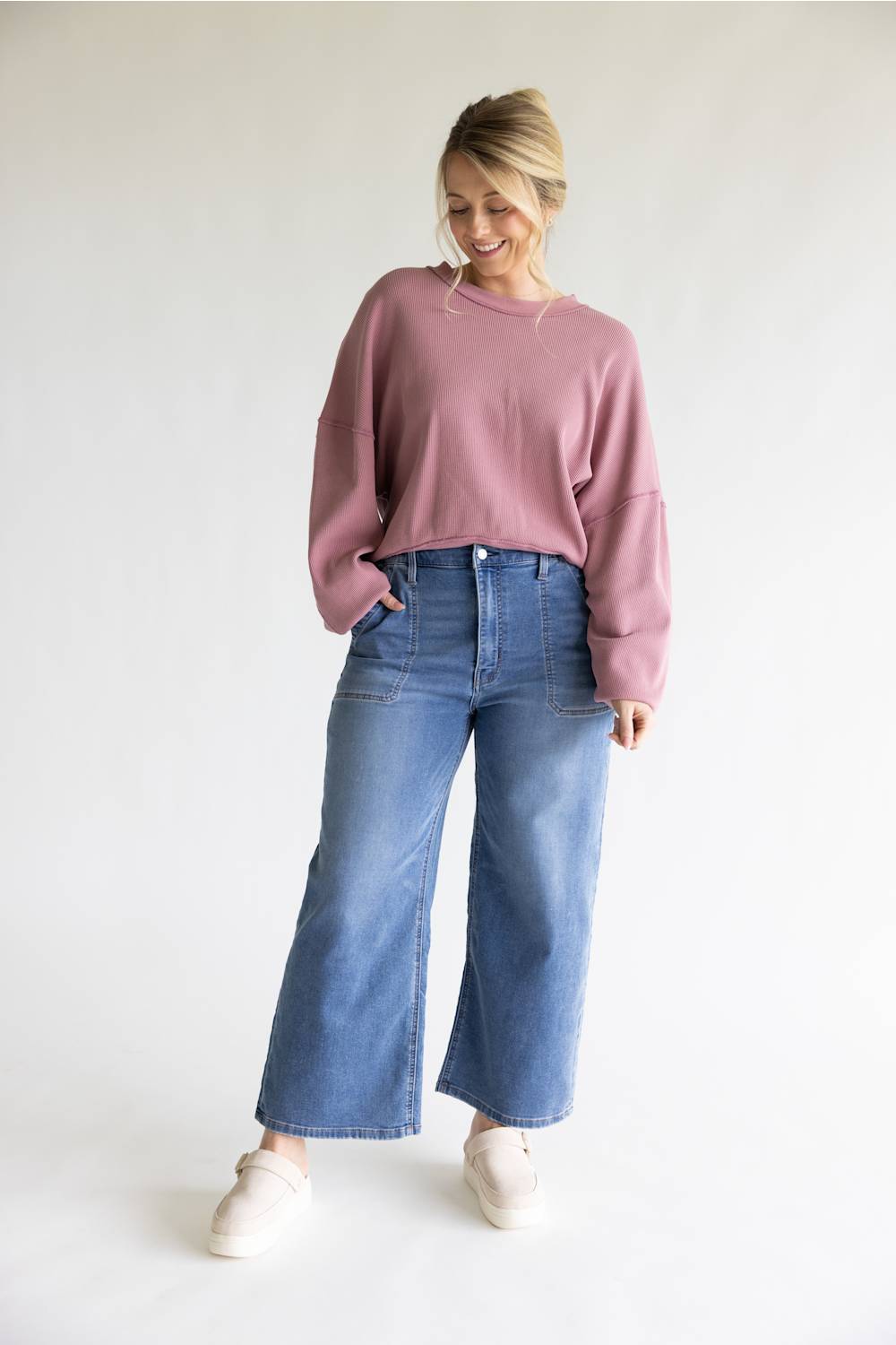 A woman modeling a dusty rose long-sleeve cropped sweatshirt and wide-leg blue jeans with white slip-on shoes, standing against a plain white studio background.