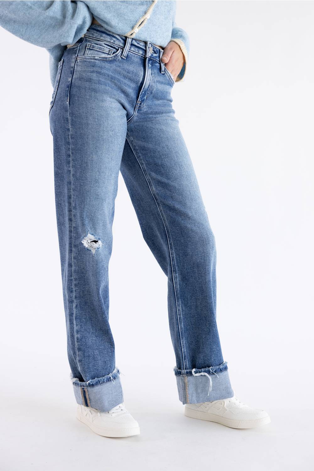 A person wearing light blue distressed jeans with rolled cuffs and white sneakers, shown from the waist down against a clean white studio background.