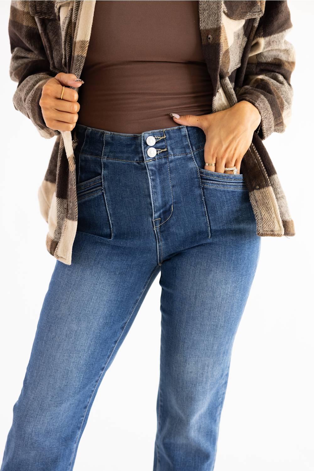 A person wearing blue high-waisted jeans with a brown bodysuit and a brown, beige, and black plaid flannel shirt, shown from the chest down against a white background.