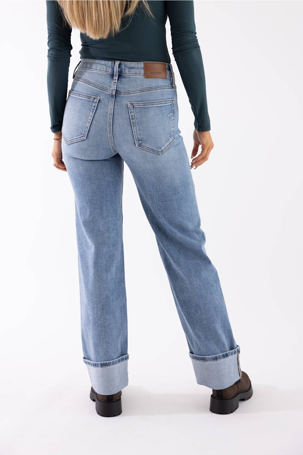 A person shown from the back wearing light-wash blue straight-leg jeans with rolled cuffs, a dark teal long-sleeve top, and dark brown shoes against a plain white studio background.