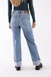 A person shown from the back wearing light-wash blue straight-leg jeans with rolled cuffs, a dark teal long-sleeve top, and dark brown shoes against a plain white studio background.