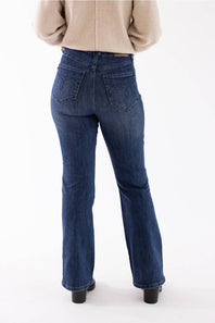 Rear view of a person wearing dark blue flared denim jeans with a beige sweater and black shoes, standing against a plain white studio background.
