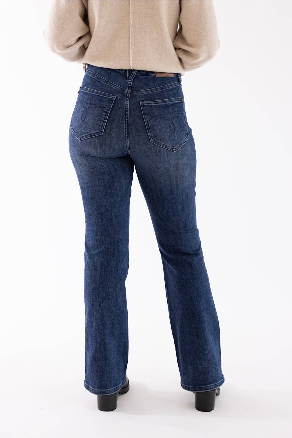 Rear view of a person wearing dark blue flared denim jeans with a beige sweater and black shoes, standing against a plain white studio background.
