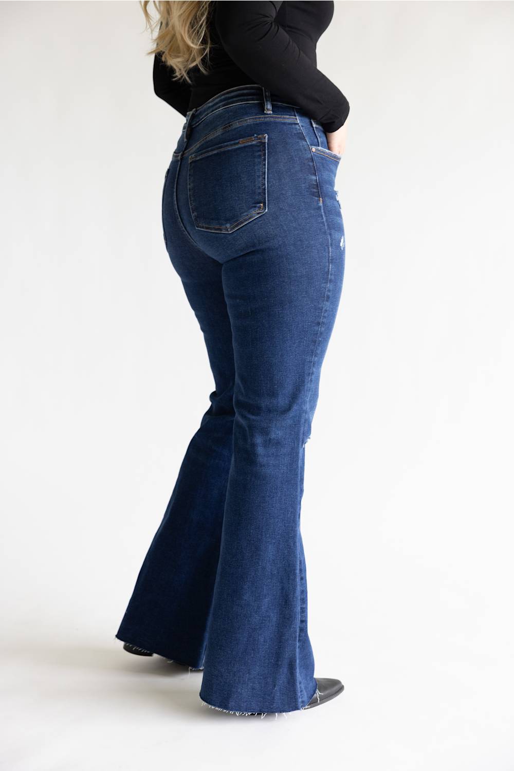 A person shown from the back wearing dark blue high-waisted flare jeans and a black long-sleeve top, standing against a plain light gray studio background.