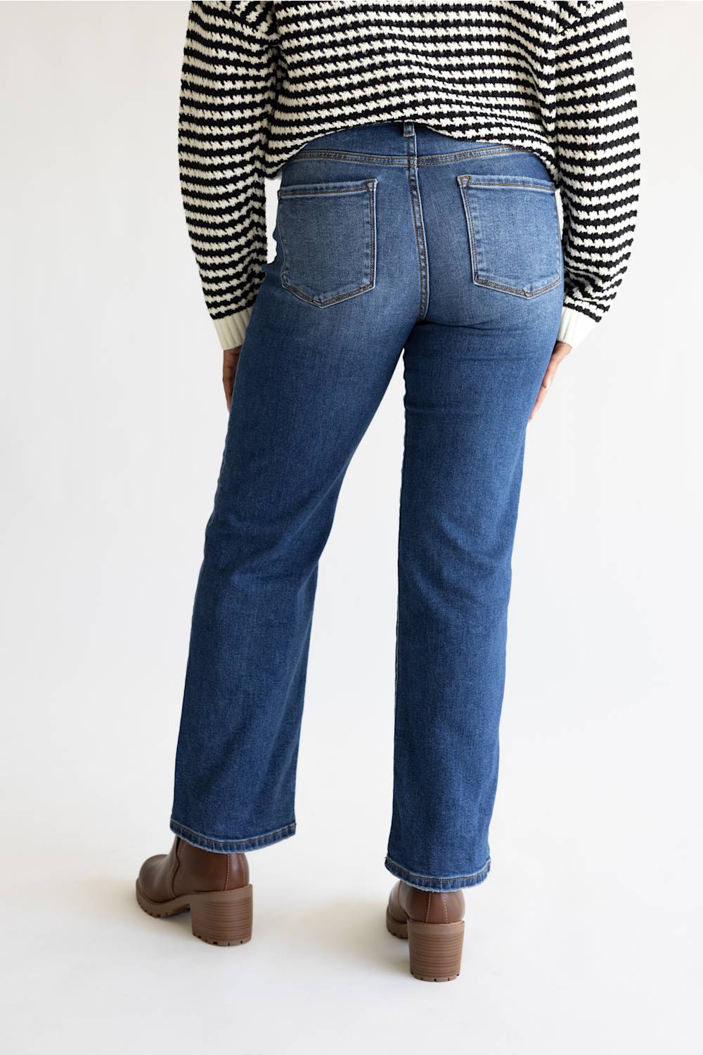 Rear view of a person wearing blue straight-leg jeans, a black and white patterned sweater, and brown block-heel boots against a plain white studio background.