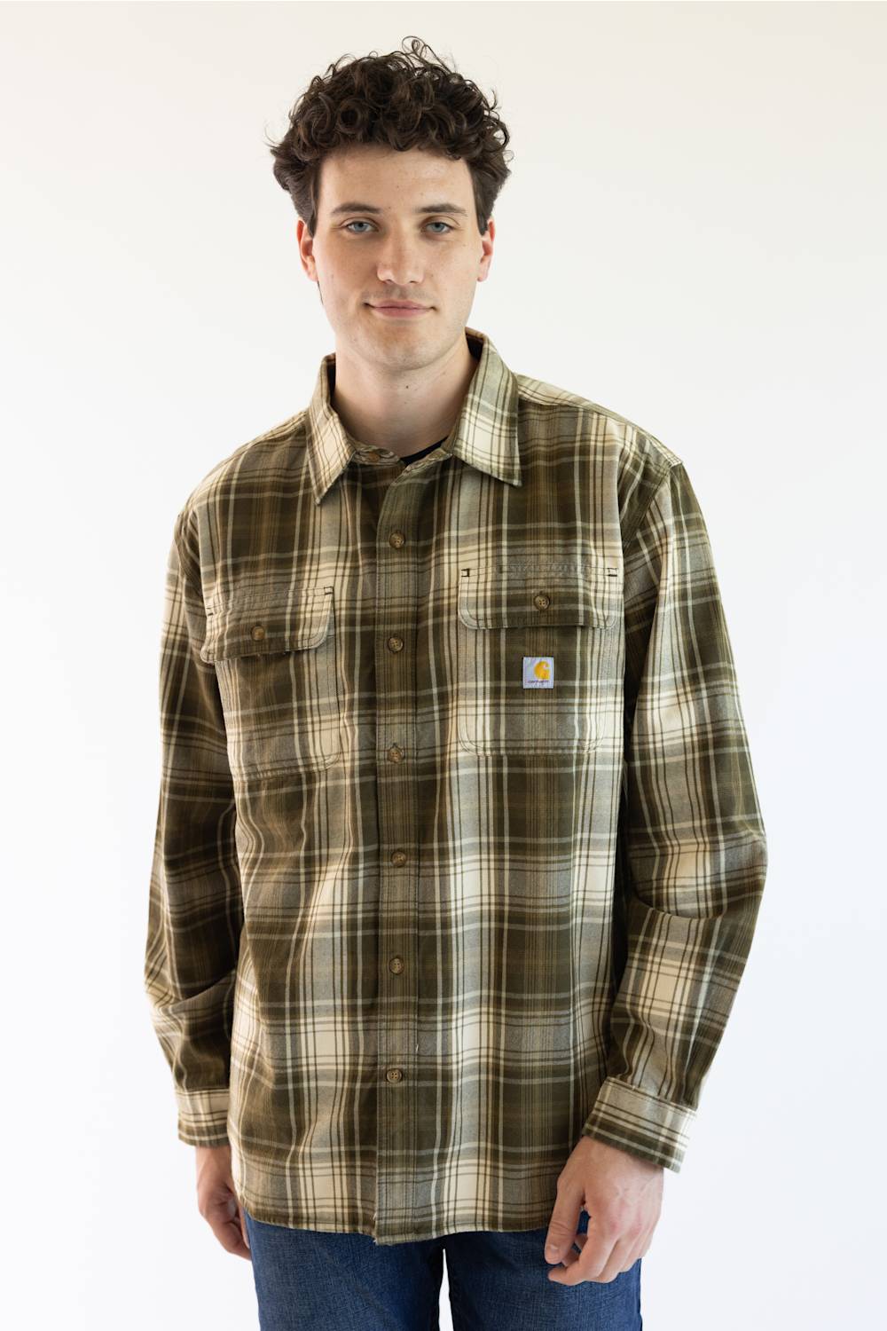 A man wearing a Carhartt long-sleeve plaid button-up shirt in olive green, brown, and beige, paired with blue jeans, photographed against a plain white studio background.
