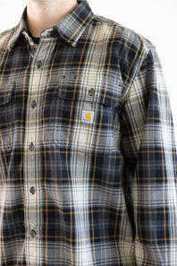 A close-up of a Carhartt plaid button-up shirt worn by a model. The shirt features a navy, gray, and beige plaid pattern with thin yellow lines, and the signature yellow Carhartt logo patch is visible on the left chest pocket.