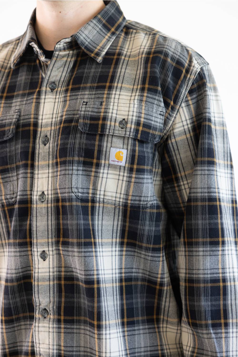 A close-up of a Carhartt plaid button-up shirt worn by a model. The shirt features a navy, gray, and beige plaid pattern with thin yellow lines, and the signature yellow Carhartt logo patch is visible on the left chest pocket.