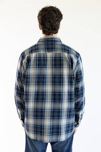 A man viewed from behind wearing a long-sleeve plaid button-up shirt in navy, blue, and beige, paired with blue jeans against a plain white background.