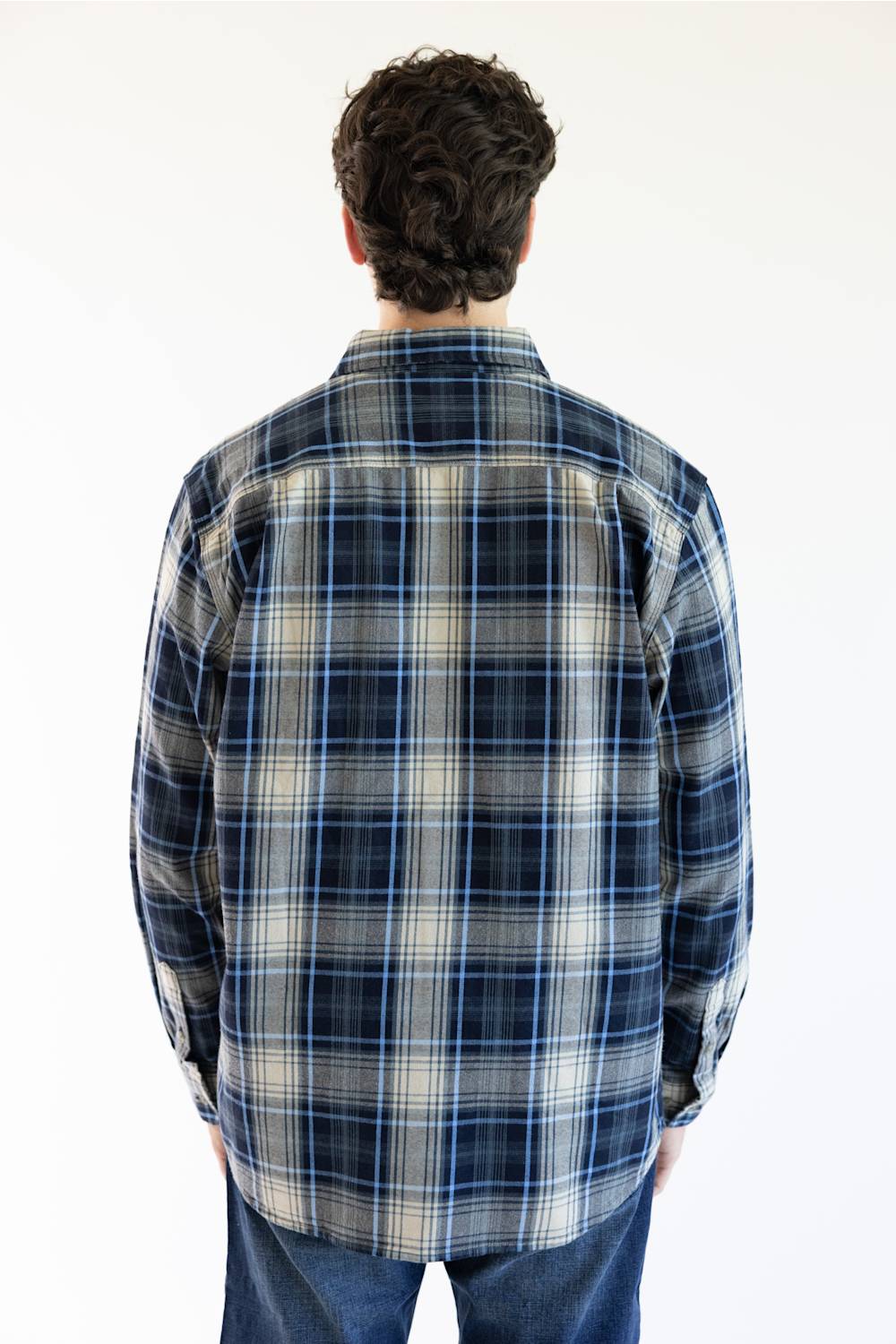 A man viewed from behind wearing a long-sleeve plaid button-up shirt in navy, blue, and beige, paired with blue jeans against a plain white background.