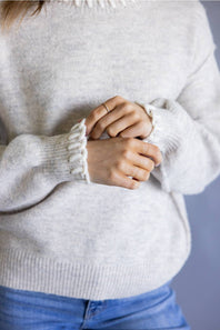 A close-up of a person wearing a light heather gray knit sweater with textured cream ribbed cuffs and a ruffled neckline, paired with blue jeans.