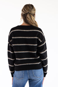 Rear view of a model wearing a black crew neck sweater with tan and white embroidered horizontal stripes across the back and long sleeves, paired with blue denim jeans.