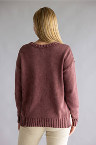 A woman viewed from behind wearing a dusty rose knit sweater and cream pants against a plain gray studio background.