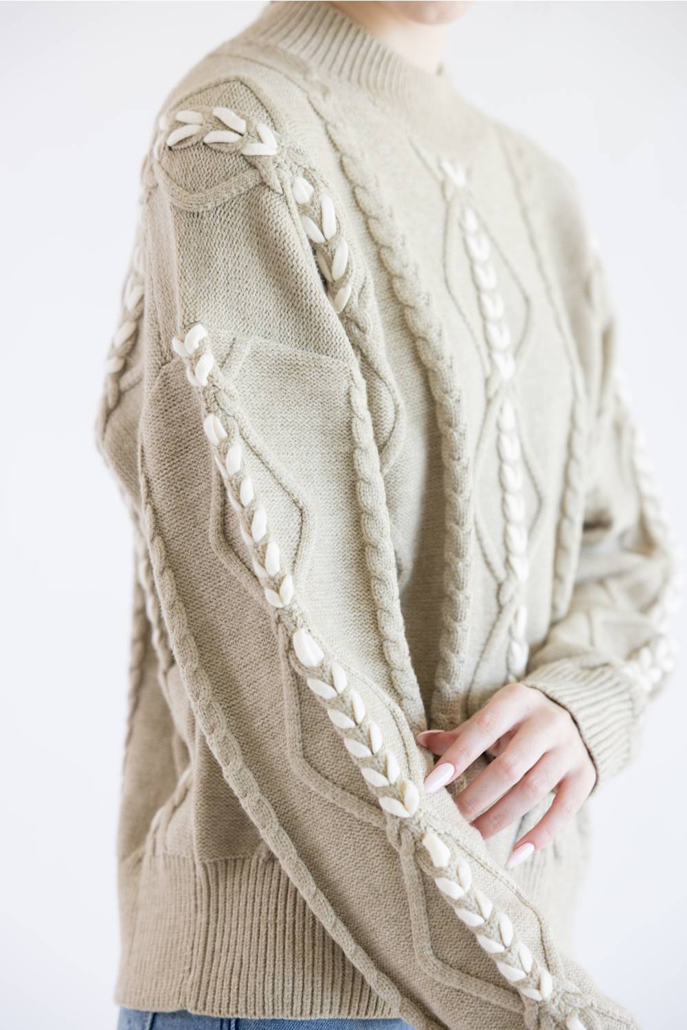 A close-up of a person wearing a beige cable-knit sweater with prominent white braided yarn detailing running down the sleeves and shoulders, set against a plain white background.