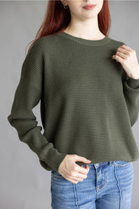 A woman wearing a dark olive green waffle-knit long-sleeve sweater and blue jeans, shown from the chin down against a plain gray studio background.
