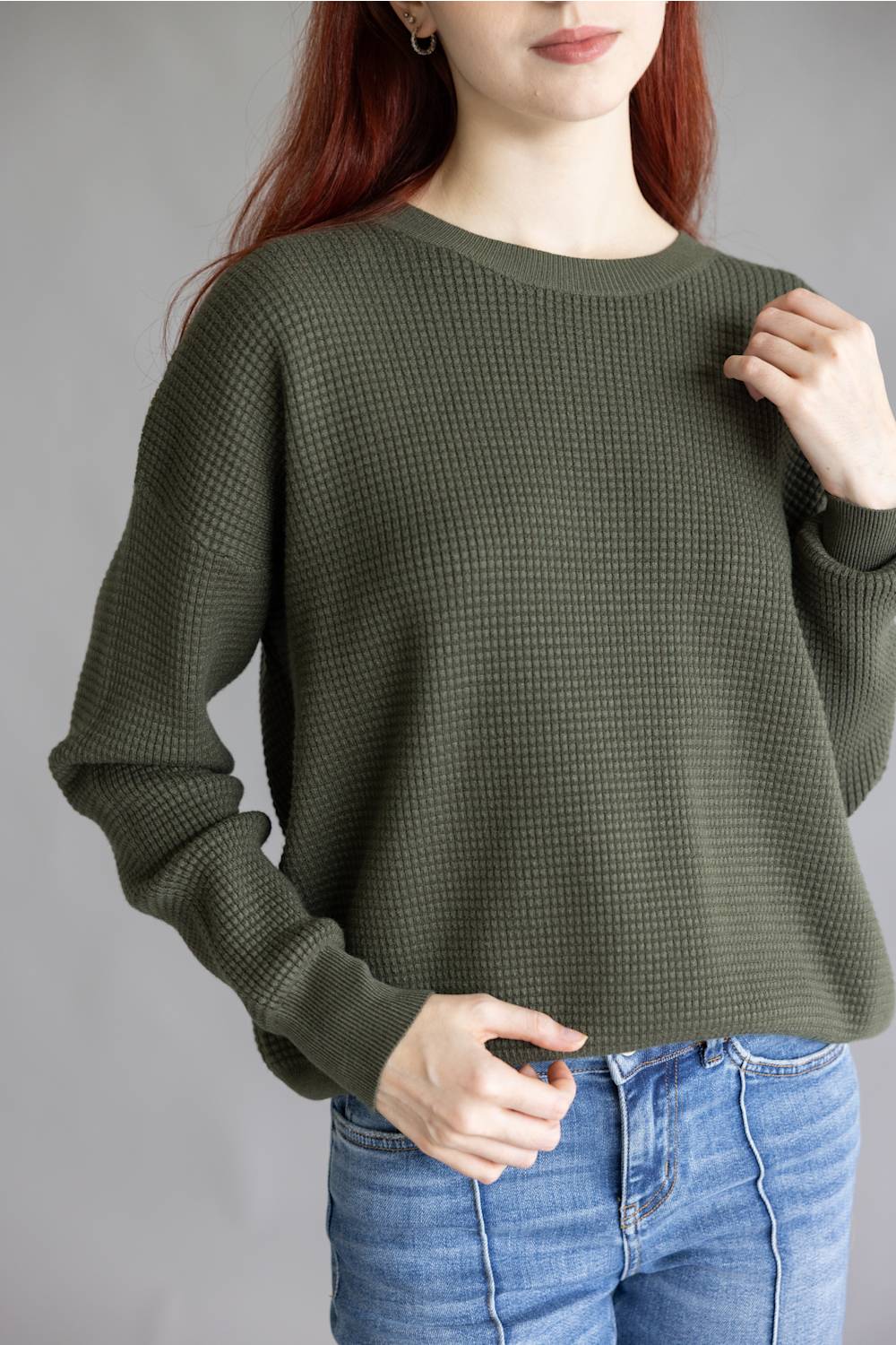 A woman wearing a dark olive green waffle-knit long-sleeve sweater and blue jeans, shown from the chin down against a plain gray studio background.