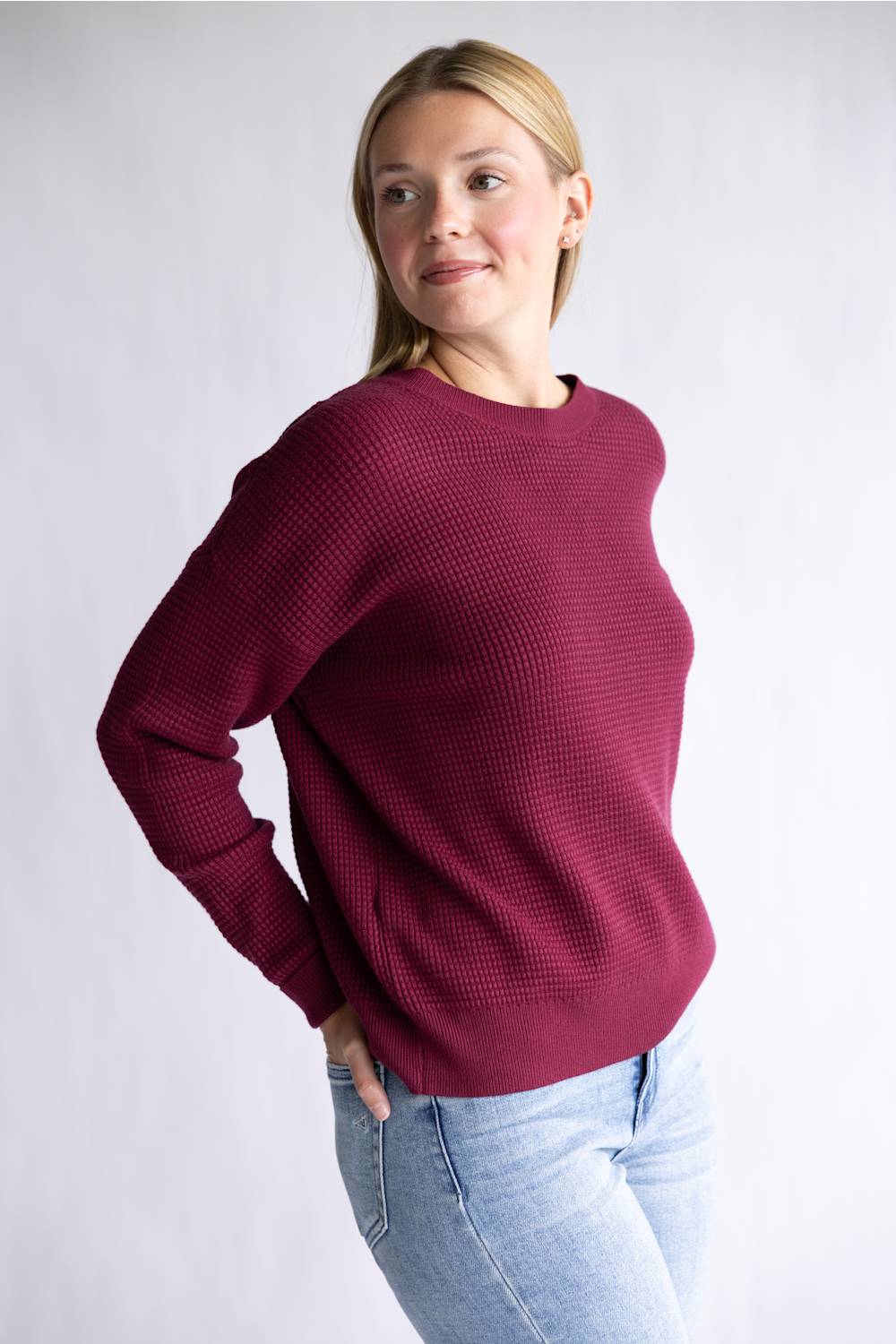 A woman wearing a textured waffle-knit crewneck sweater in deep burgundy, paired with light blue jeans, standing against a light gray studio background.