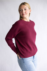 A woman wearing a textured waffle-knit crewneck sweater in deep burgundy, paired with light blue jeans, standing against a light gray studio background.