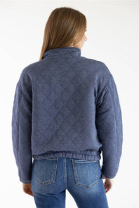 A woman viewed from the back wearing a quilted blue jacket and blue jeans against a plain white studio background.