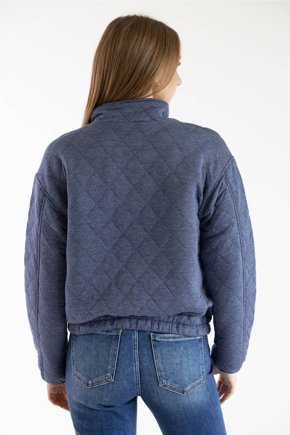 A woman viewed from the back wearing a quilted blue jacket and blue jeans against a plain white studio background.