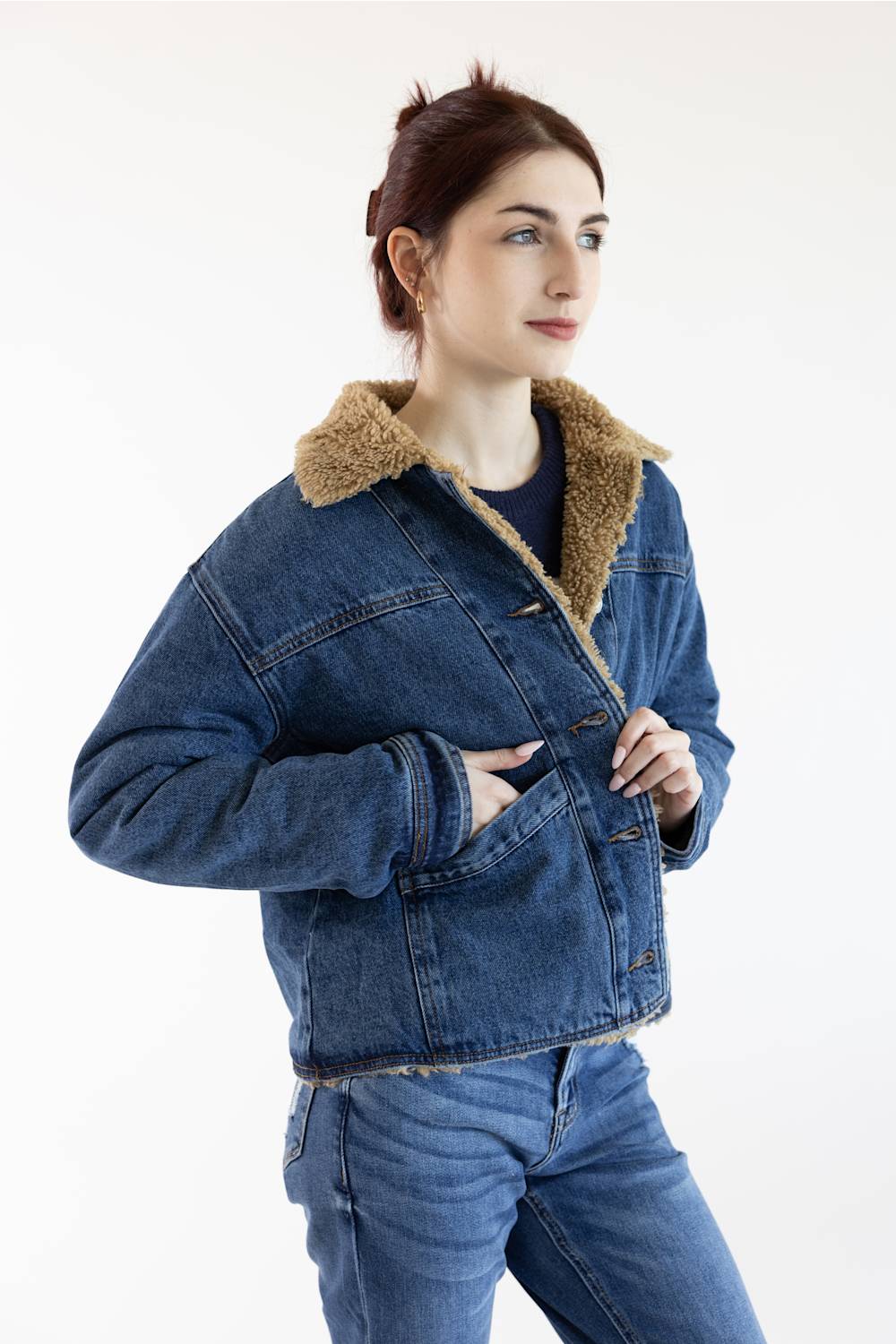 A woman wearing a cropped blue denim jacket with a tan sherpa fleece collar, paired with blue jeans, photographed against a plain white studio background.