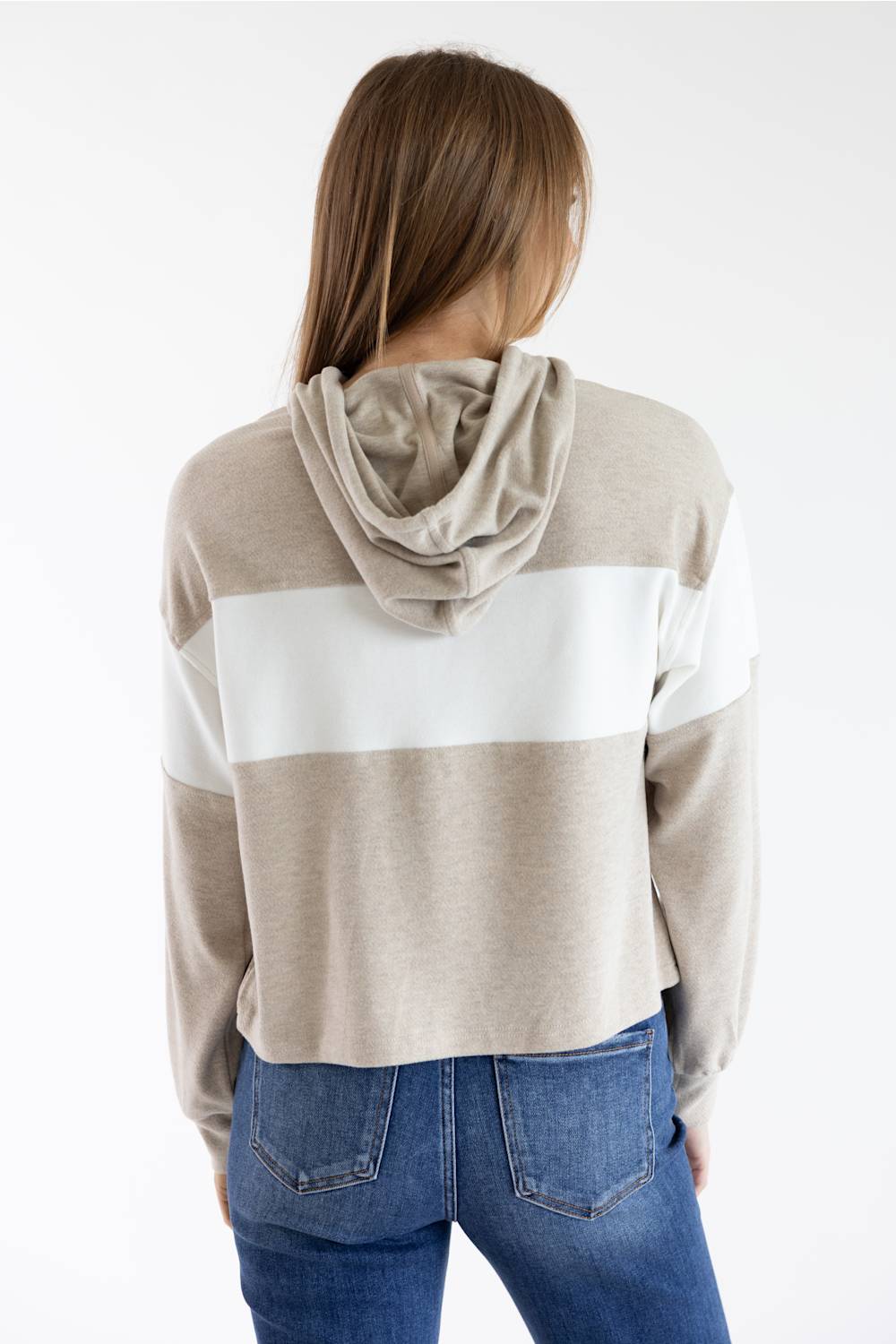 A woman shown from the back wearing a cropped hooded sweatshirt with horizontal beige and white color blocking, paired with blue jeans, against a plain white studio background.
