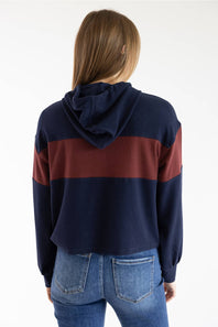 A woman viewed from behind wearing a cropped navy blue hoodie with a wide burgundy stripe across the shoulders and upper back, paired with blue jeans, against a plain white background.