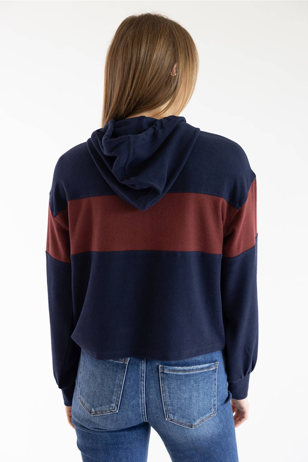 A woman viewed from behind wearing a cropped navy blue hoodie with a wide burgundy stripe across the shoulders and upper back, paired with blue jeans, against a plain white background.