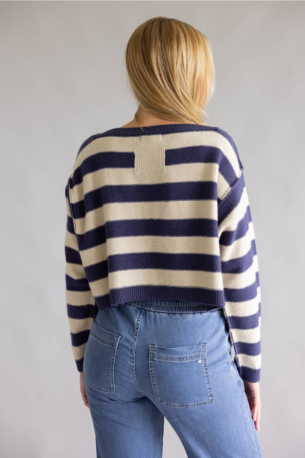 A woman viewed from behind wearing a navy and cream striped knit sweater and light blue jeans against a plain light gray studio background.