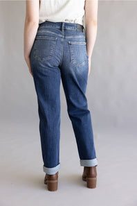 A person shown from the back wearing medium-wash blue jeans with rolled cuffs and brown block-heel shoes against a plain light gray studio background.