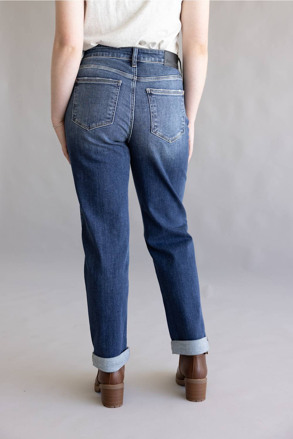 A person shown from the back wearing medium-wash blue jeans with rolled cuffs and brown block-heel shoes against a plain light gray studio background.