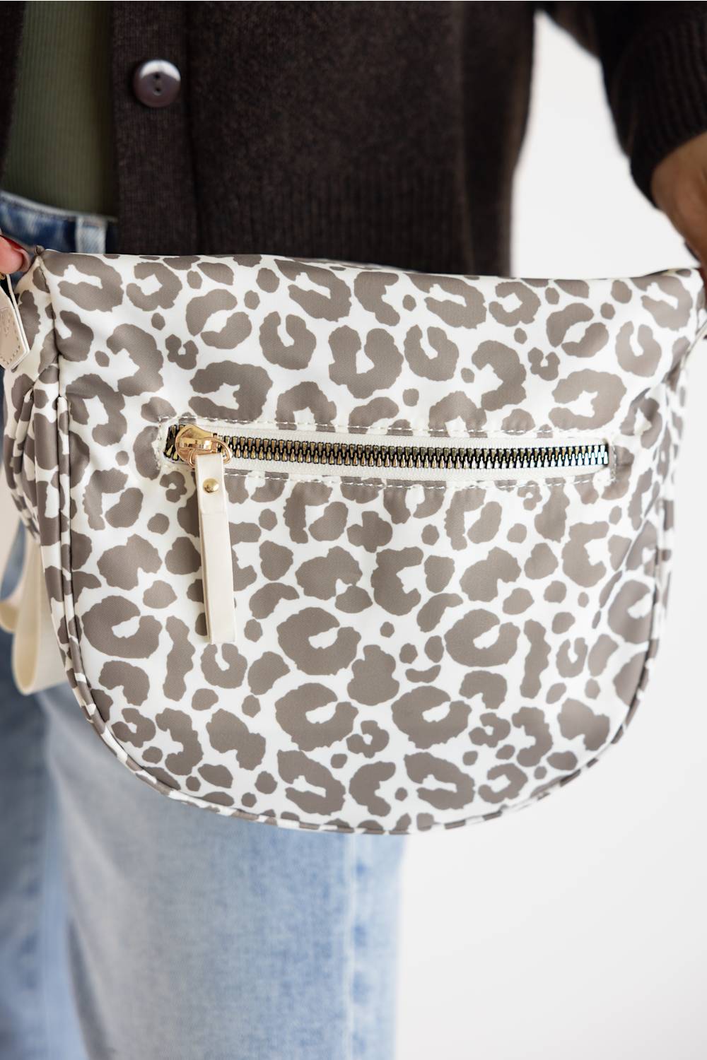A close-up of a person wearing a leopard print fanny pack. The bag features a white base with brown and gray leopard spots, a front zippered pocket with a gold-tone zipper pull, and is shown being worn with a dark brown cardigan and light blue jeans.