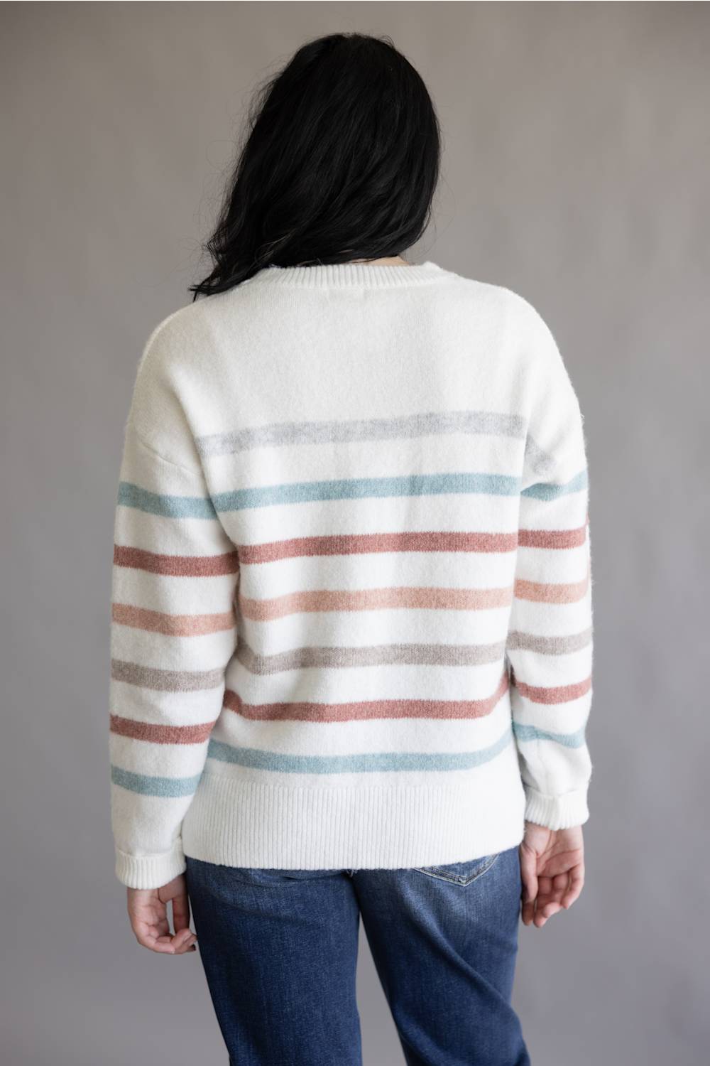 A person shown from the back wearing a cream knit sweater with horizontal dusty pink, light blue, and gray stripes, paired with dark blue jeans, against a plain gray background.